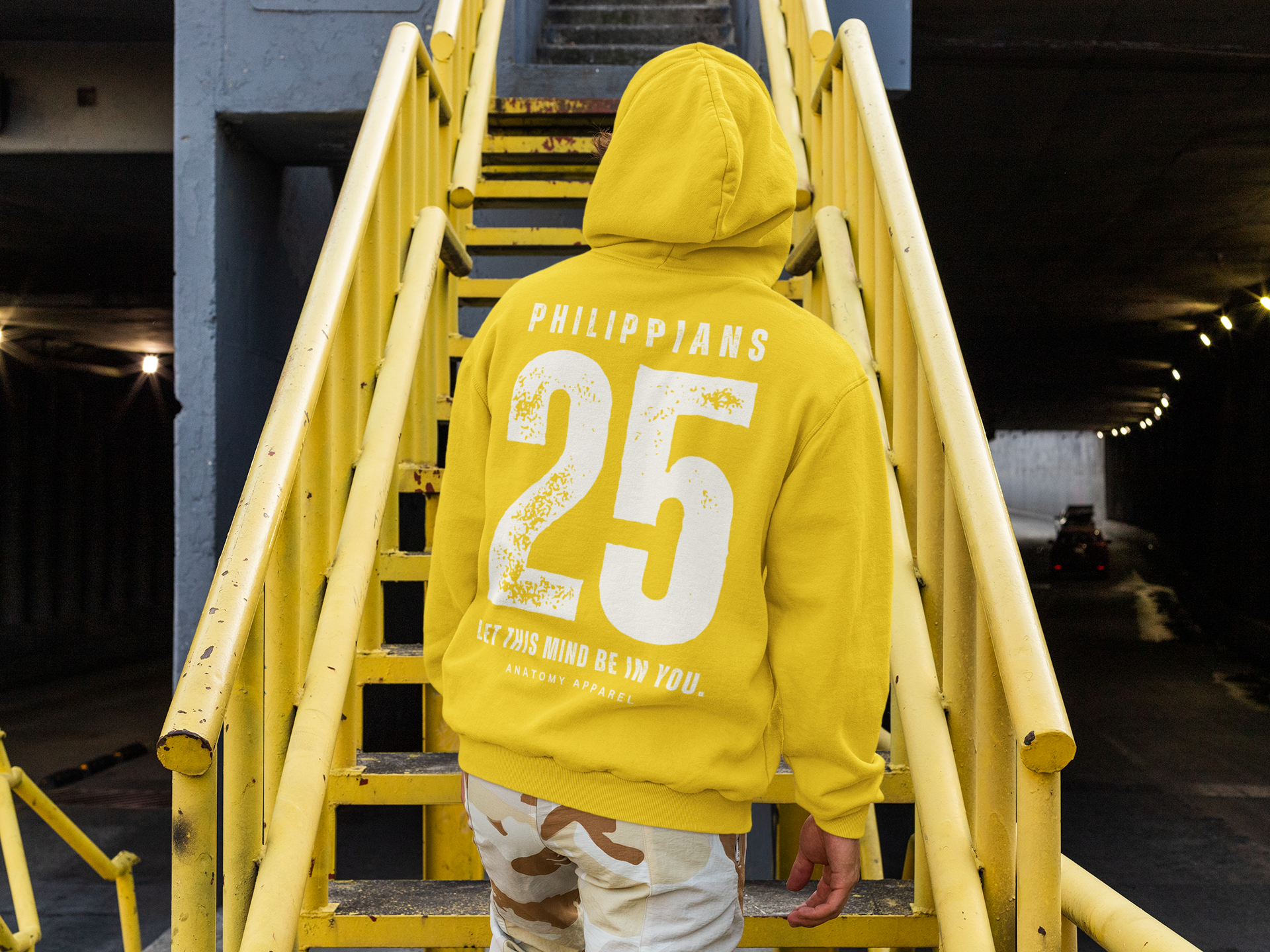 Anatomy Apparel man wearing a yellow hoodie with 'Philippians 2:5' text on a yellow staircase.