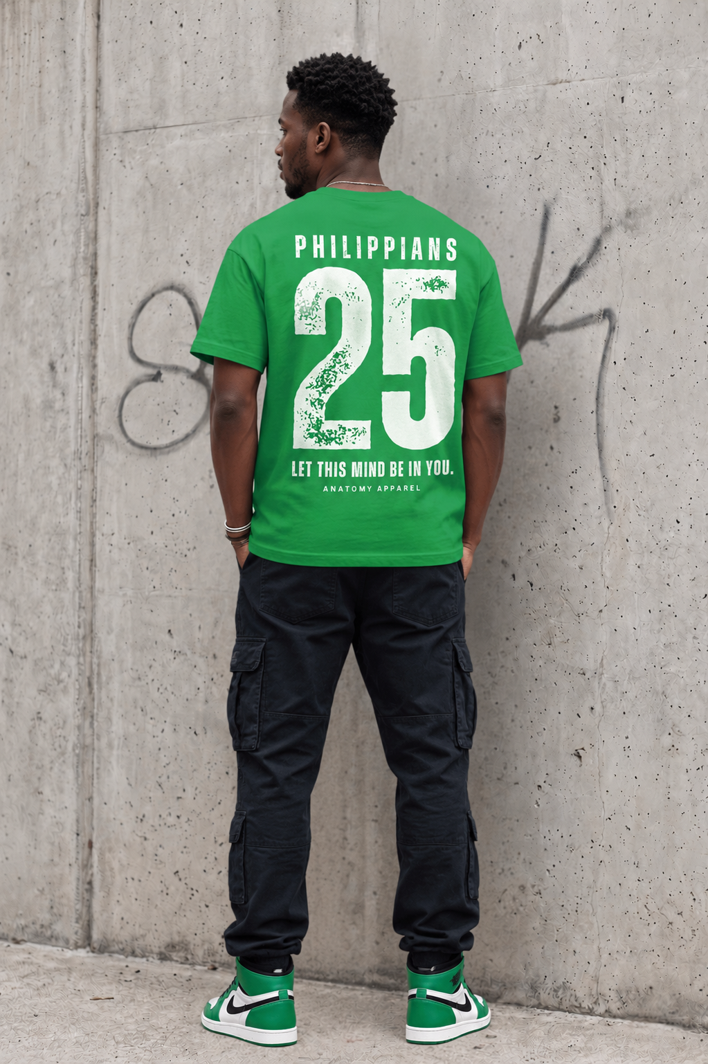 Person wearing a green t-shirt with 'Philippians 2:5' text against a concrete wall.